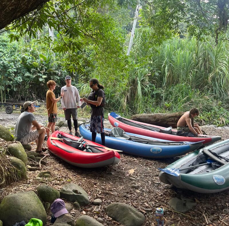 The image shows a group of people gathered around several inflatable kayaks on a riverbank. The kayaks are red, blue, and green. The people appear to be preparing for a kayaking trip. They are standing near the kayaks, possibly getting ready to launch them into the water. The surrounding environment includes lush greenery and trees, suggesting a natural setting.